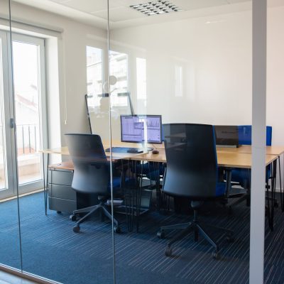 Empty boardroom behind glass wall. Meeting room with conference table, shared desk for team and workplaces. Trading graphs on monitor. Office interior or commercial real estate concept